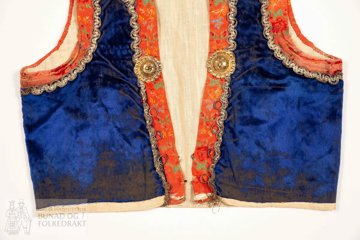 Liv, lapp med "brudeliv 1908".
Liv i blå silkeplysj/langhåra fløyel på bomullsbott kanta med 3,3 cm breie raude silkeband med blomemønstring i raudt, gult, blått og grønt. Innafor ei 1,5 cm agramanblonde.
Sidesaum og skuldersaum er bakoverflytta, rett ringing i hals bak, med snippa som peika nedover. Mjukt forma framkant som møtest i midja.
I framkant, på kvar side, fire maljer festa i stoffet mellom silkeband og agramanblonda, med belegg i bomullslerret på bakside. Første malje er 3,2 cm opp frå nedkant, siste 12,7 cm opp.
19 cm frå nedkanten på det raude silkebandet er ei stor bore (4 cm bred). Løv mangla.
På innsida ein glissen, naturfarga bomullslerret. Kanta rundt til framsida.
Vrangsydd fôr i side- og skuldersauma; maskinsauma.
Raudt band er maskinsauma i ytterkant; ellers er livet berre handsauma.
I livhøgde to små, sekundære hemper og hekter.