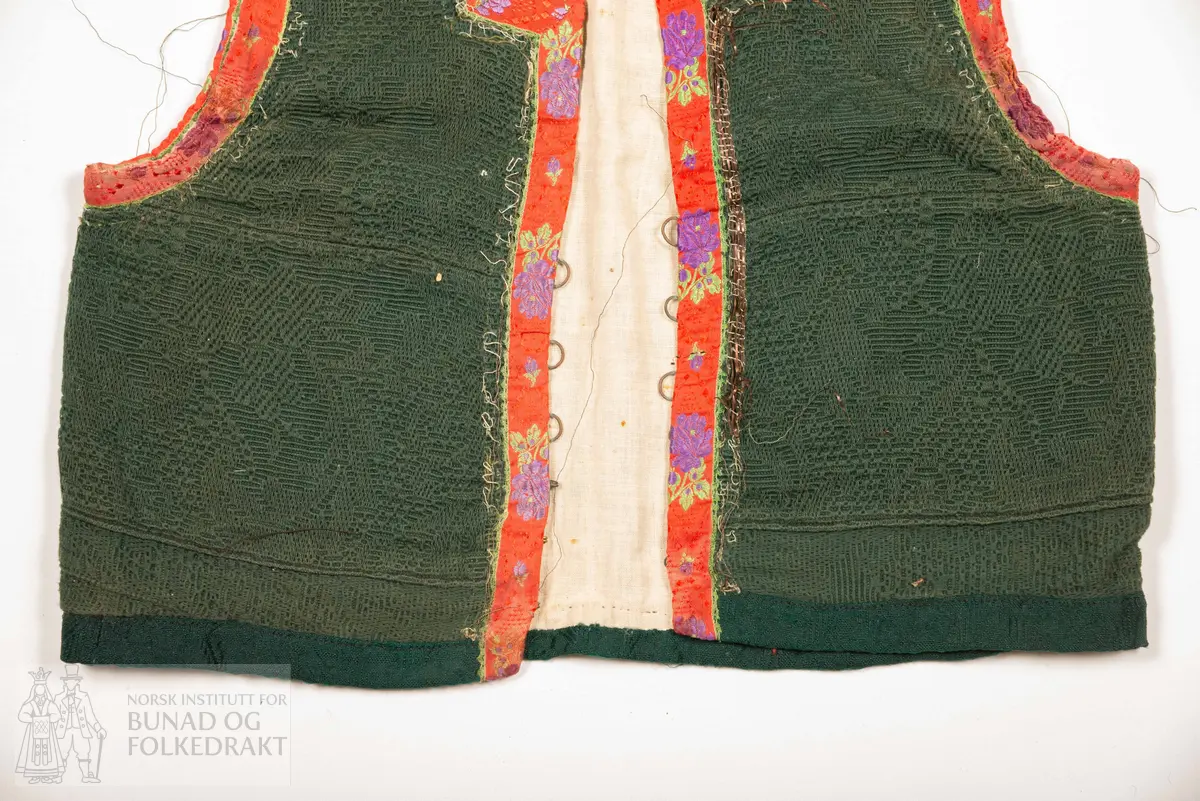 Grønt liv i jaquardvove bomullskvalitet, kanta med raude, jaquardvove silkeband med blomar i lilla og grønt.
Livet har ingen skuldersaum, og litt bakoverflytta, rette sidesauma.
Lett runda halsringing bak, rett, halvdjup utringing foran med rette hjørne som peika veldig svakt oppover.
Mjukt forma framkant som møtest nede i midja foran.
Livet er skøyta på i nedkant fram.
Resta etter eit 0,6 cm metallband innafor 1,7 cm bredt silkeband.
Kanta nede med grøn amiens.
På kvar side i framkant er det tre hekter og hemper (første 2,8 cm opp, siste 7,7 cm), og over dei er tre majer (9,6 cm opp, siste 15,6 cm).
Fôra med tynn glissen bomullslerret. Ei ekstra remse under hekte og hempe, og under malje.
Sidesaumane er jarekanter.
Handsauma.