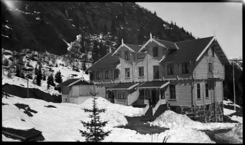 Oversiktsbilde over Krokan hotells bakside. Sanitærbygg for hotellets gjester på byggets gavlvegg mot nord. To kvinner stående på trappa.