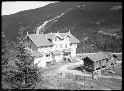 Oversikt over baksiden av Krokan hotell med to laftede stabbur. Foss i fjellet i bakgrunnen i forbindelse med spyling av rør for Norsk Hydro. Barn sittende på trappa til hotellet og hund gående på gårdsplassen. Sanitærbygg på hotellets nordside og torvtak med pipe synlig i bildets forgrunn.