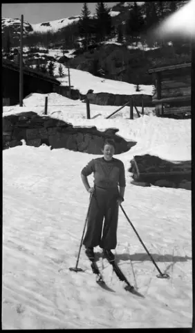 Kvinne på ski på Krokan. Stabbur og laftet bolighus for Fossogårdene i bakgrunnen.