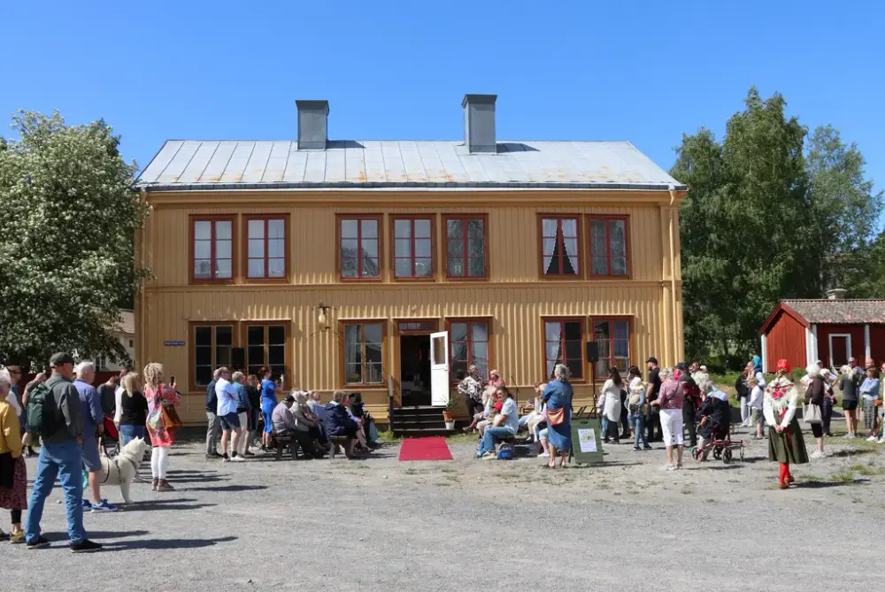 Ett gult hus med dörren öppen och en skara folk som sitter och står utanför.