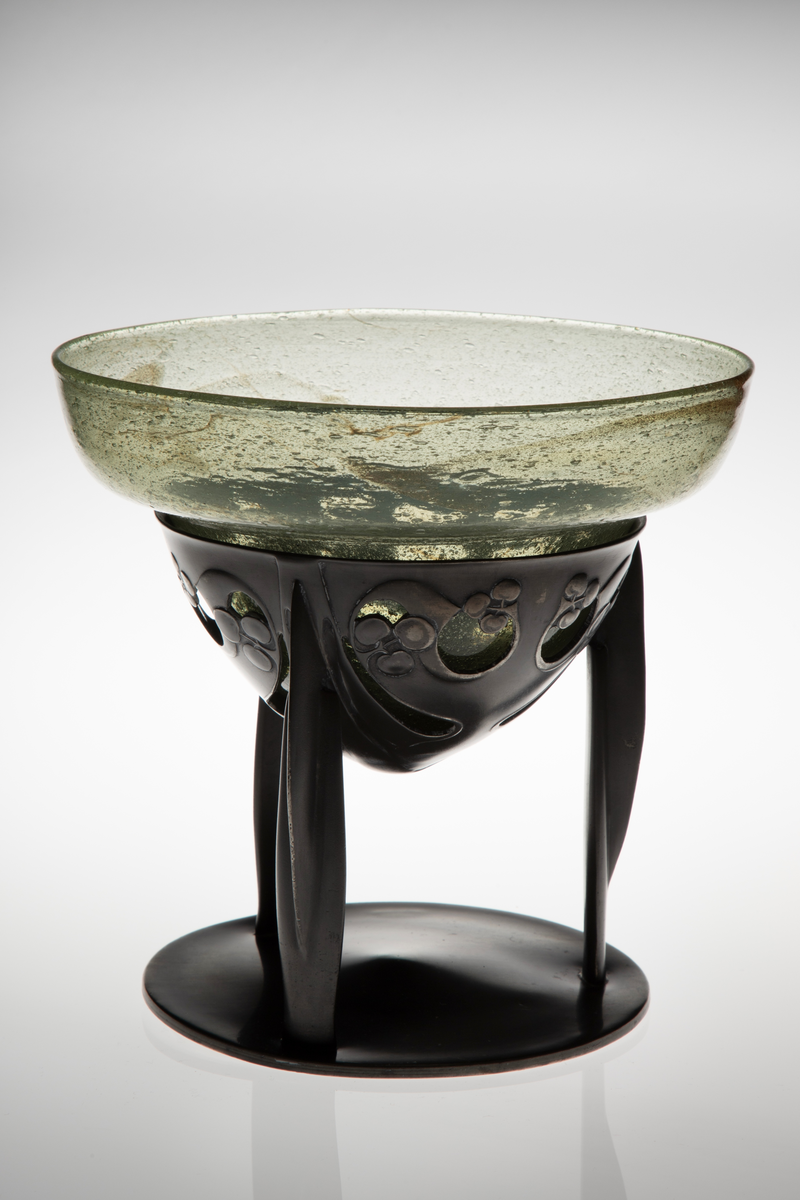 Glasskål som hviler på trebent tinnfot. Skålen er klokkeformet med bred kant, og den eplegrønne glassmassen er fylt med små blærer. Langs kanten er det partier med innlagt bladgull. Den nedre delen bæres av en trebent fot i tinn, hvis overflate er oksidert. Den øvre delen er gjennombrutt, og smykket med stiliserte planteornamenter i lavt relieff. De tre benene hviler på sirkulær fotplate, som er noe hevet mot midten.