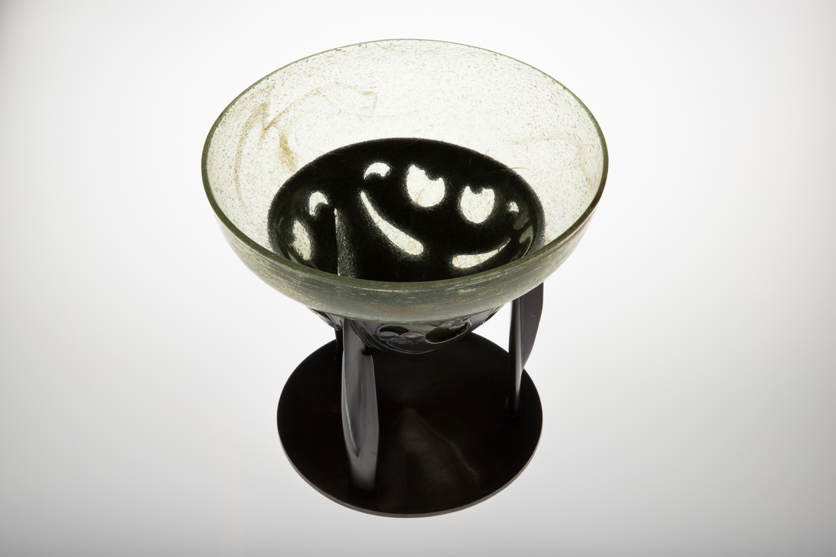 Glasskål som hviler på trebent tinnfot. Skålen er klokkeformet med bred kant, og den eplegrønne glassmassen er fylt med små blærer. Langs kanten er det partier med innlagt bladgull. Den nedre delen bæres av en trebent fot i tinn, hvis overflate er oksidert. Den øvre delen er gjennombrutt, og smykket med stiliserte planteornamenter i lavt relieff. De tre benene hviler på sirkulær fotplate, som er noe hevet mot midten.