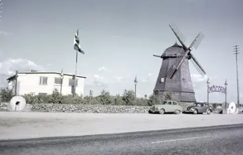 Exteriörbild. Restaurang Möllan i Morup. Vid kvarnen drevs både boende och restaurang. Entrén mellan lyktstolparna flankeras av kvarnhjul och där står två bilar parkerade; de ena är en "folkabubbla". Tvärs över bilden går telefonledningar.