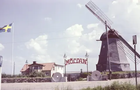 Exteriörbild. Restaurang Möllan i Morup. Här drevs både boende och restaurang. Entrén mellan lyktstolparna flankeras av kvarnhjul. I bildens förgrund finns en busshållplatsskylt till höger och en parkeringsskylt till vänster.