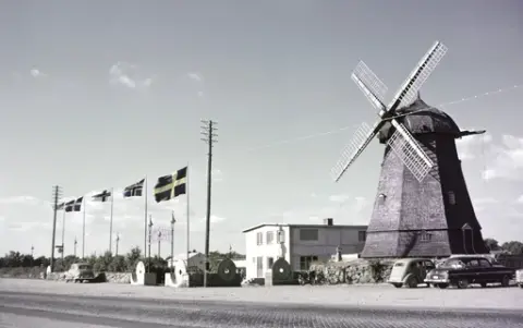 Exteriörbild. Restaurang Möllan i Morup. Vid kvarnen drevs både boende och restaurang. Entrén mellan lyktstolparna flankeras av kvarnhjul och fyra länders flaggor är hissade. Några bilar står parkerade under el- och telefonledningarna som går utmed vägen.