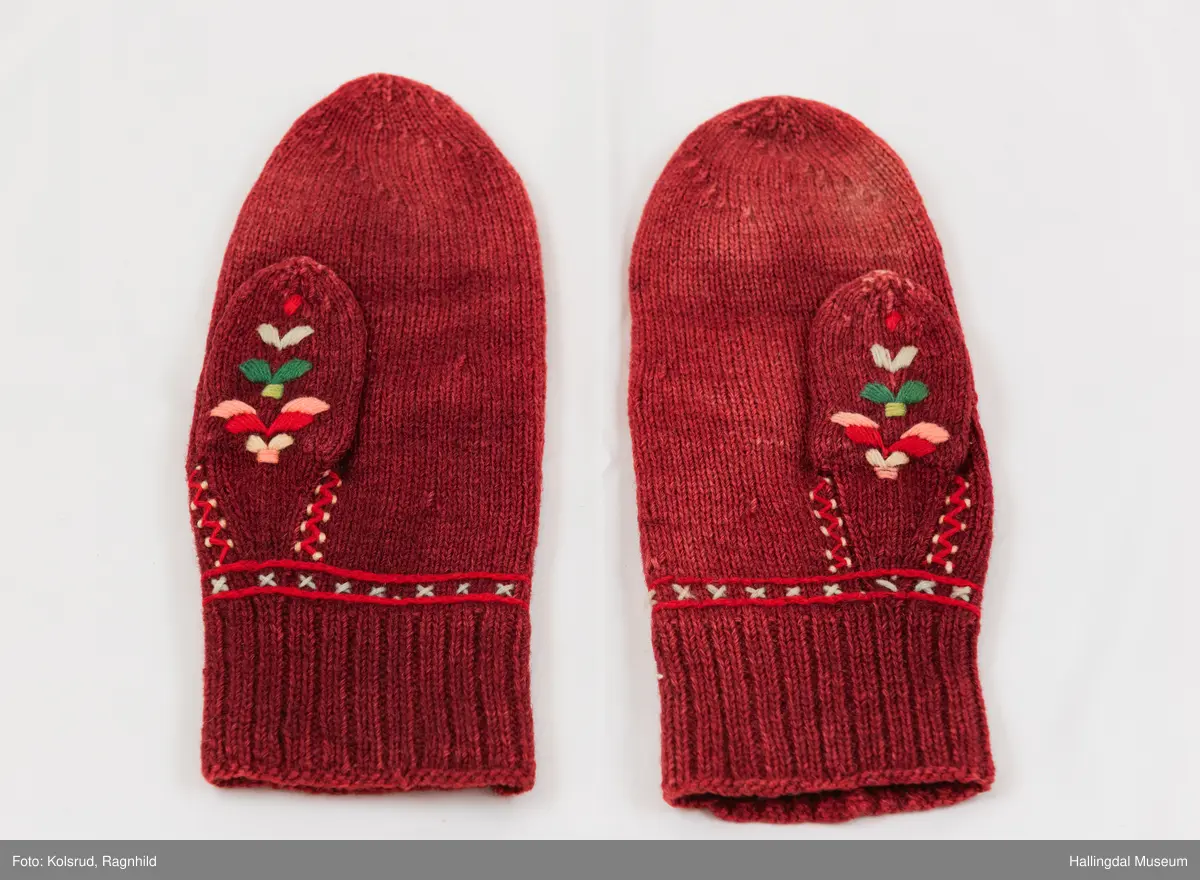 Votter strikka av raudbrunt ullgarn. Stilisert blomstremotiv på framsida, i tjukksaum med flanillsgarn i 3 raudfargar, raudorange, gult, 2 grønfargar og kvitt.