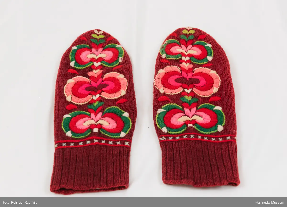 Votter strikka av raudbrunt ullgarn. Stilisert blomstremotiv på framsida, i tjukksaum med flanillsgarn i 3 raudfargar, raudorange, gult, 2 grønfargar og kvitt.