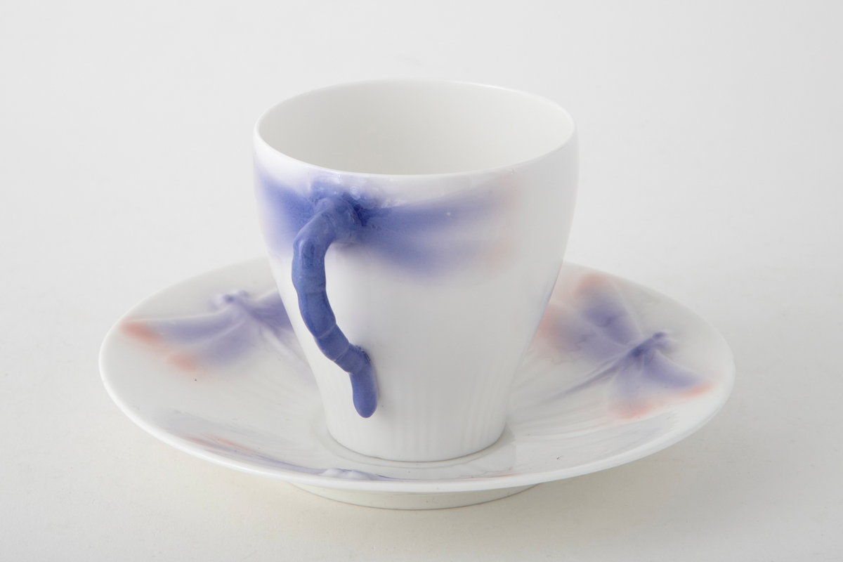 Sjablondreiet porselen. Hanken er støpt. Kopp med konisk form dekorert med plastiske øyenstikkere med rosa og blå underglasur på hvit grunn.