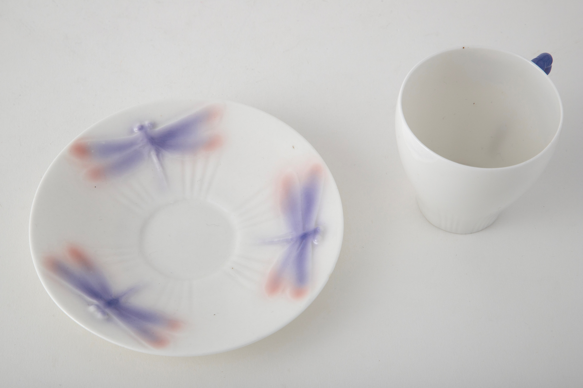 Sjablondreiet porselen. Hanken er støpt. Kopp med konisk form dekorert med plastiske øyenstikkere med rosa og blå underglasur på hvit grunn.