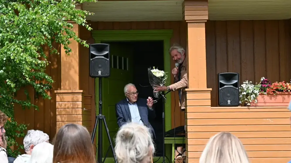 Museumsleiar ved Vinjesenteret Yngve Knausgård overrekk Olav Vesaas ein blomsterbukett under opninga av sommarsesongen på Midtbø. Olav sit og Yngve står på trappa på hovudhuset på Midtbø.