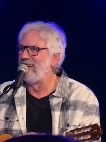 Louis Jacoby, mann med grått hår, grått skjegg og briller holder gitar og synger inn i mikrofon. Han smiler. Bakgrunnen er mørk blå.