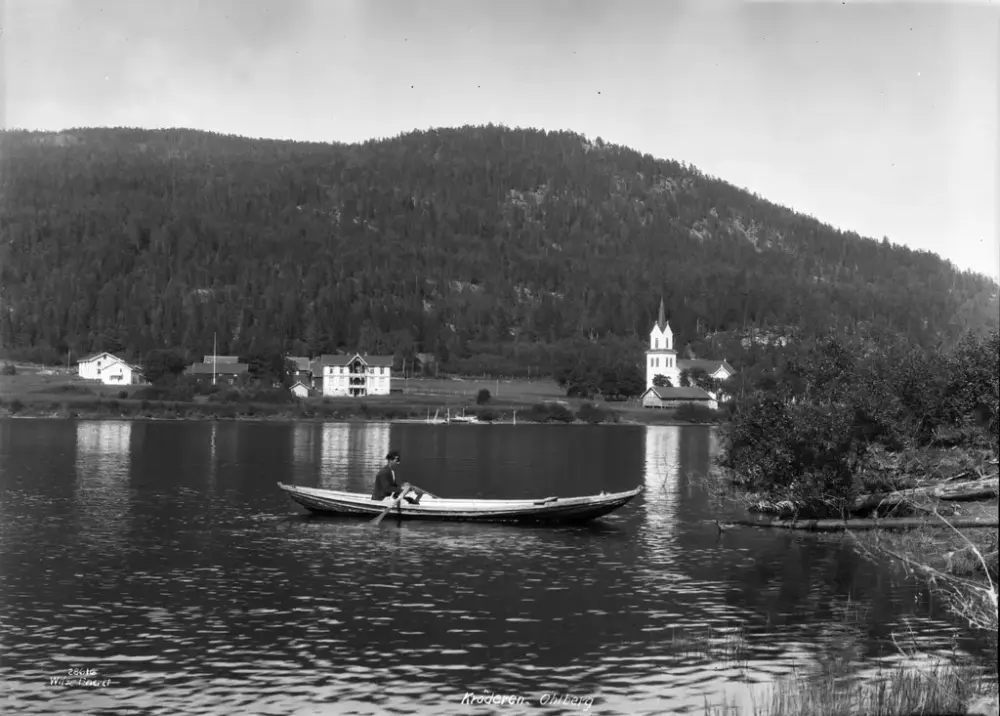 Svart-hvitt foto av en mann i robåt på Krøderfjorden. I bakgrunnen er Sparebankgården og Olberg kirke.