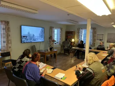 Personer sitter runt ett halvcikelformat bord och tittar på en skärm, och en annan person som talar framför skärmen.