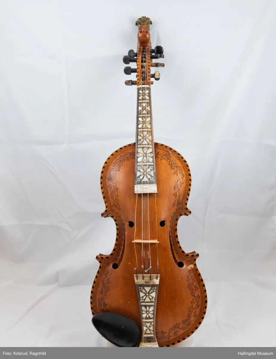 Hardingfele. Fela er litt kuven på midten, og har to S-forma lydhol i loket. Gripebrettet er dekorert med innfelte bitar av perlemor, og det er skore eit dyrehovud med krone i enden av halsen. Fela er dekorert langs kanten, med blomedekor. 