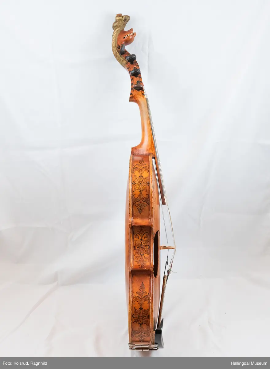 Hardingfele. Fela er litt kuven på midten, og har to S-forma lydhol i loket. Gripebrettet er dekorert med innfelte bitar av perlemor, og det er skore eit dyrehovud med krone i enden av halsen. Fela er dekorert langs kanten, med blomedekor. 