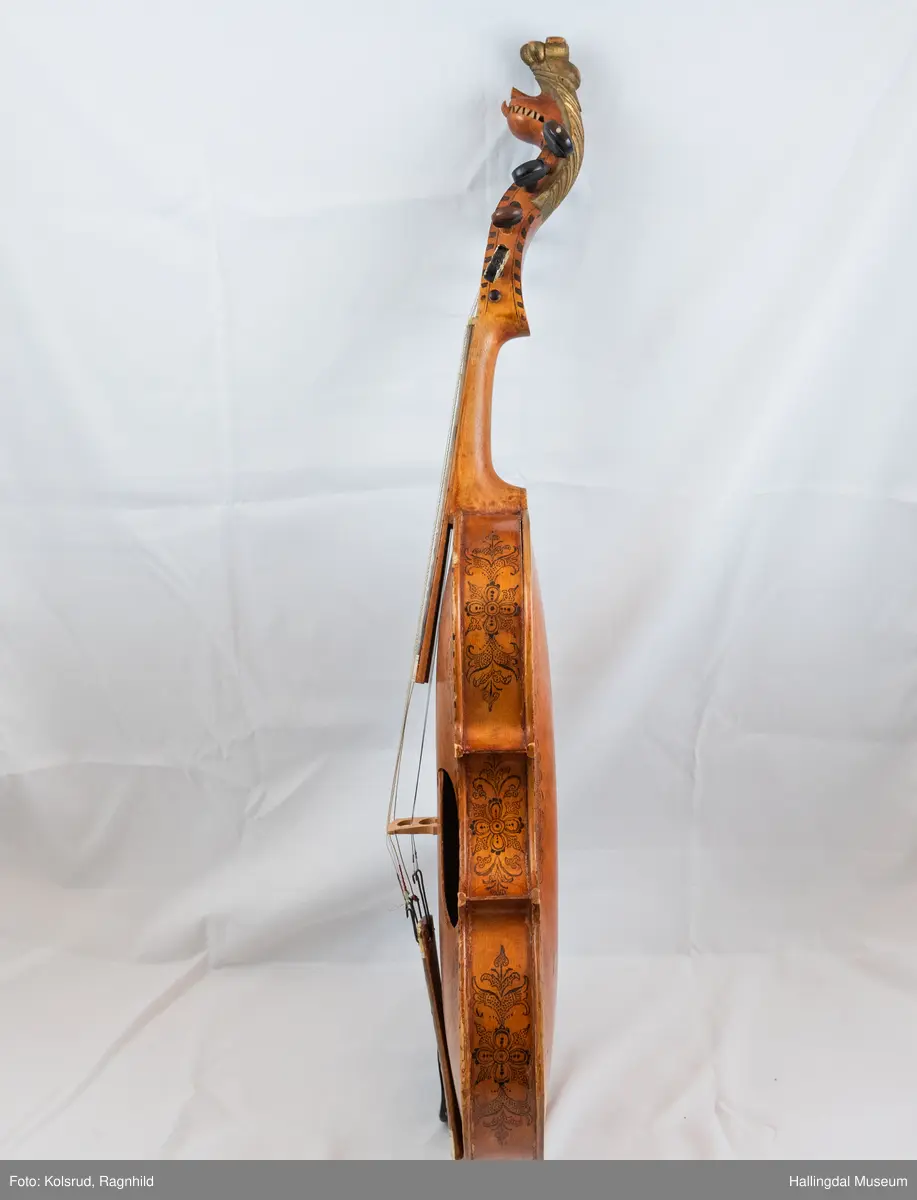 Hardingfele. Fela er litt kuven på midten, og har to S-forma lydhol i loket. Gripebrettet er dekorert med innfelte bitar av perlemor, og det er skore eit dyrehovud med krone i enden av halsen. Fela er dekorert langs kanten, med blomedekor. 