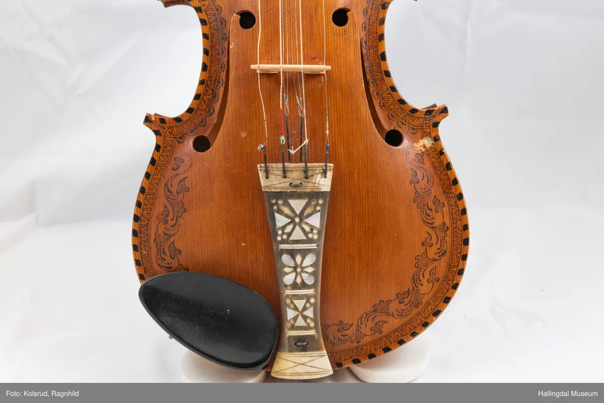 Hardingfele. Fela er litt kuven på midten, og har to S-forma lydhol i loket. Gripebrettet er dekorert med innfelte bitar av perlemor, og det er skore eit dyrehovud med krone i enden av halsen. Fela er dekorert langs kanten, med blomedekor. 