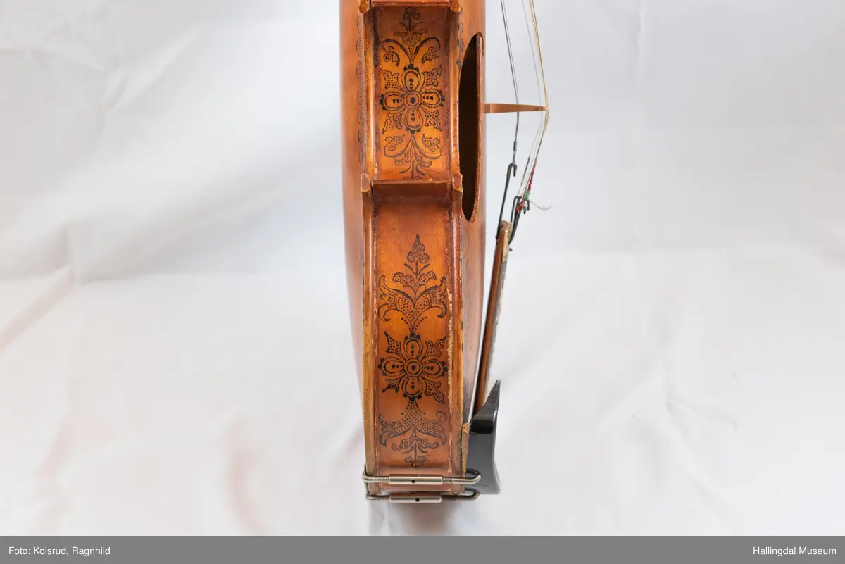 Hardingfele. Fela er litt kuven på midten, og har to S-forma lydhol i loket. Gripebrettet er dekorert med innfelte bitar av perlemor, og det er skore eit dyrehovud med krone i enden av halsen. Fela er dekorert langs kanten, med blomedekor. 