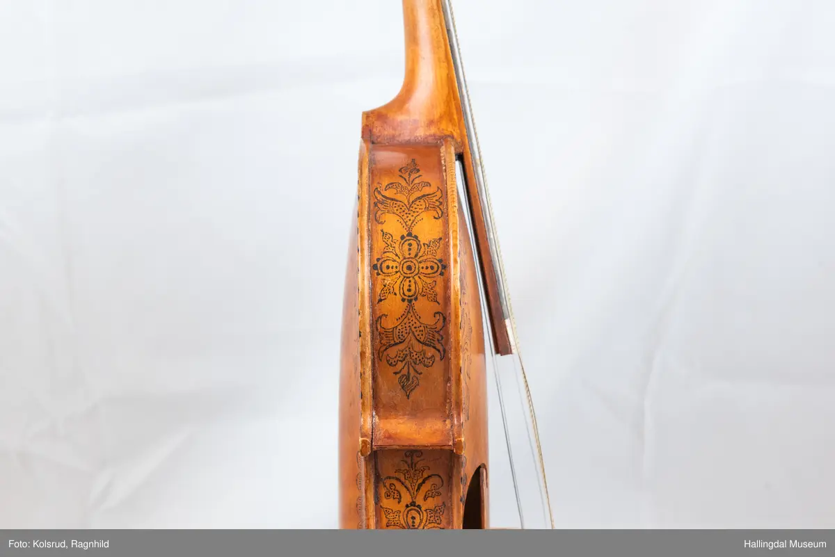 Hardingfele. Fela er litt kuven på midten, og har to S-forma lydhol i loket. Gripebrettet er dekorert med innfelte bitar av perlemor, og det er skore eit dyrehovud med krone i enden av halsen. Fela er dekorert langs kanten, med blomedekor. 