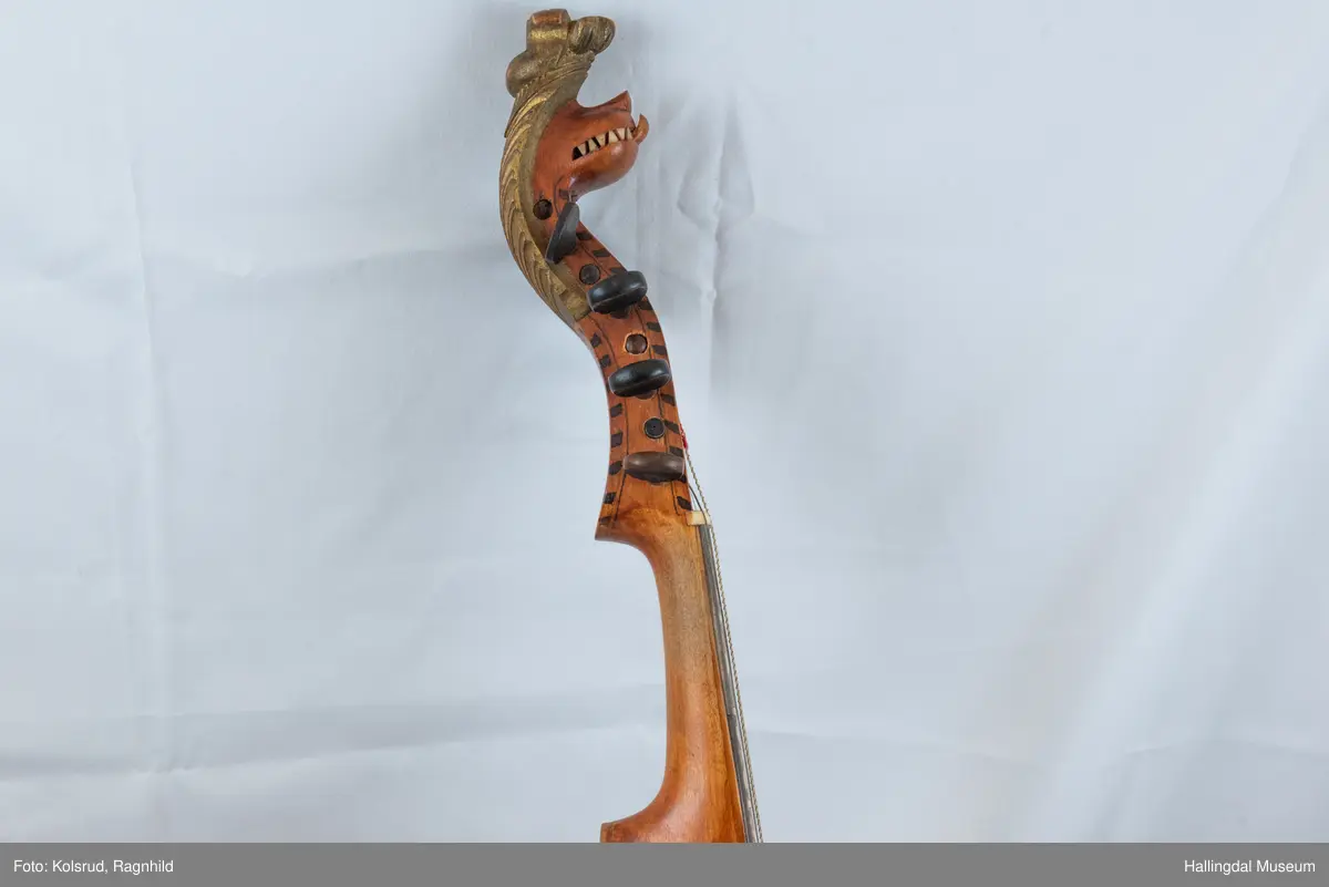 Hardingfele. Fela er litt kuven på midten, og har to S-forma lydhol i loket. Gripebrettet er dekorert med innfelte bitar av perlemor, og det er skore eit dyrehovud med krone i enden av halsen. Fela er dekorert langs kanten, med blomedekor. 