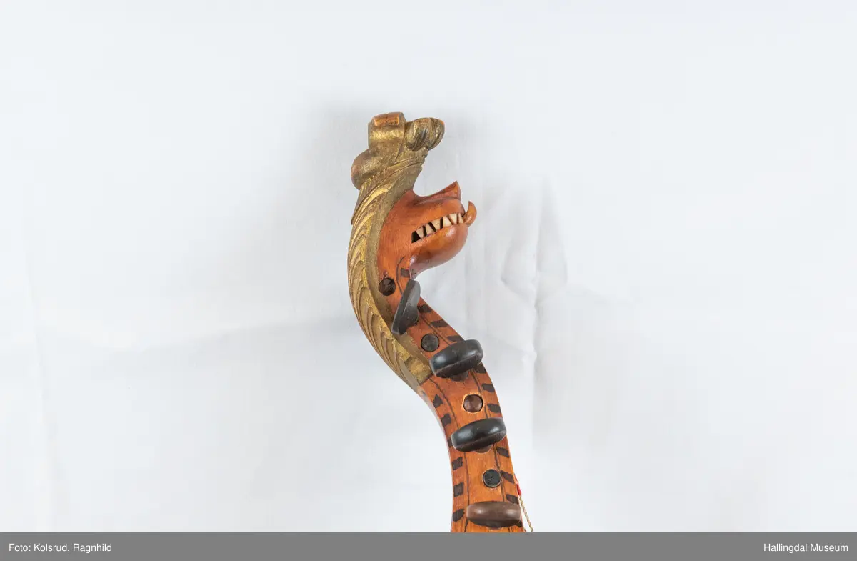 Hardingfele. Fela er litt kuven på midten, og har to S-forma lydhol i loket. Gripebrettet er dekorert med innfelte bitar av perlemor, og det er skore eit dyrehovud med krone i enden av halsen. Fela er dekorert langs kanten, med blomedekor. 