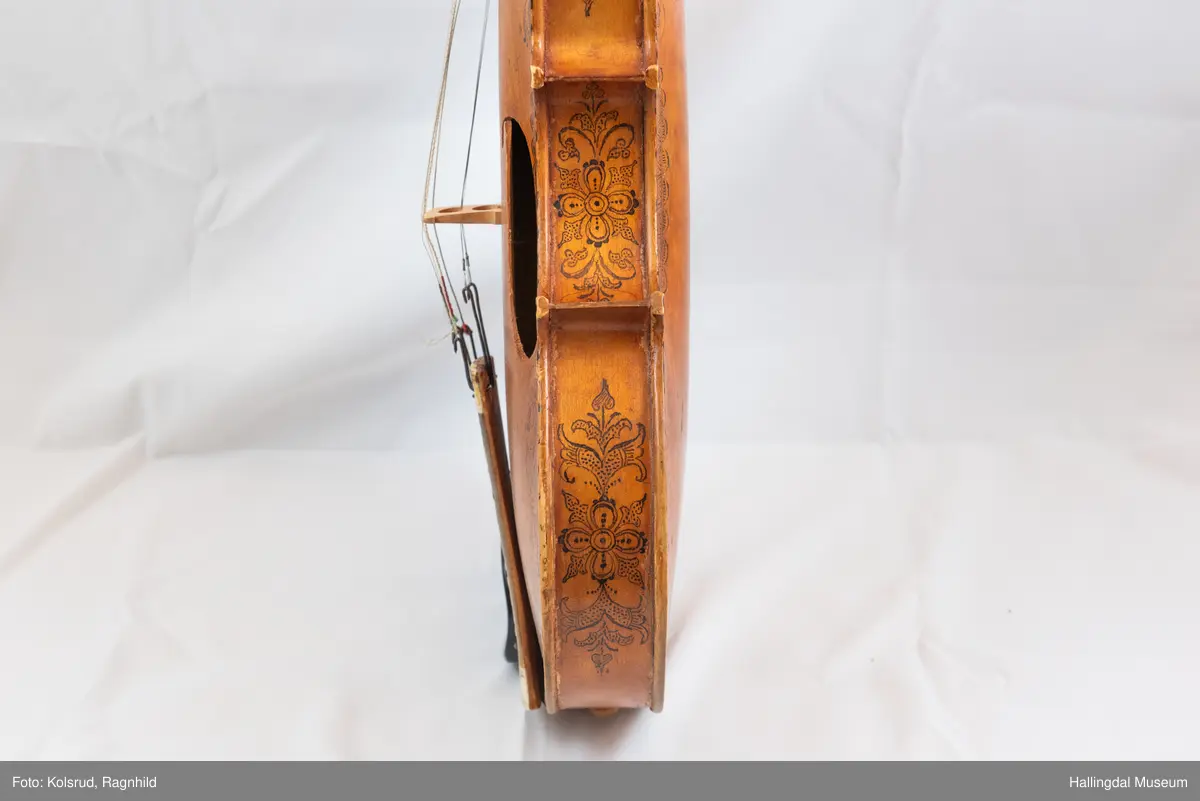 Hardingfele. Fela er litt kuven på midten, og har to S-forma lydhol i loket. Gripebrettet er dekorert med innfelte bitar av perlemor, og det er skore eit dyrehovud med krone i enden av halsen. Fela er dekorert langs kanten, med blomedekor. 