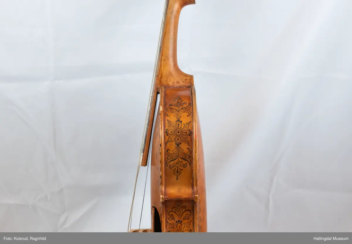 Hardingfele. Fela er litt kuven på midten, og har to S-forma lydhol i loket. Gripebrettet er dekorert med innfelte bitar av perlemor, og det er skore eit dyrehovud med krone i enden av halsen. Fela er dekorert langs kanten, med blomedekor. 