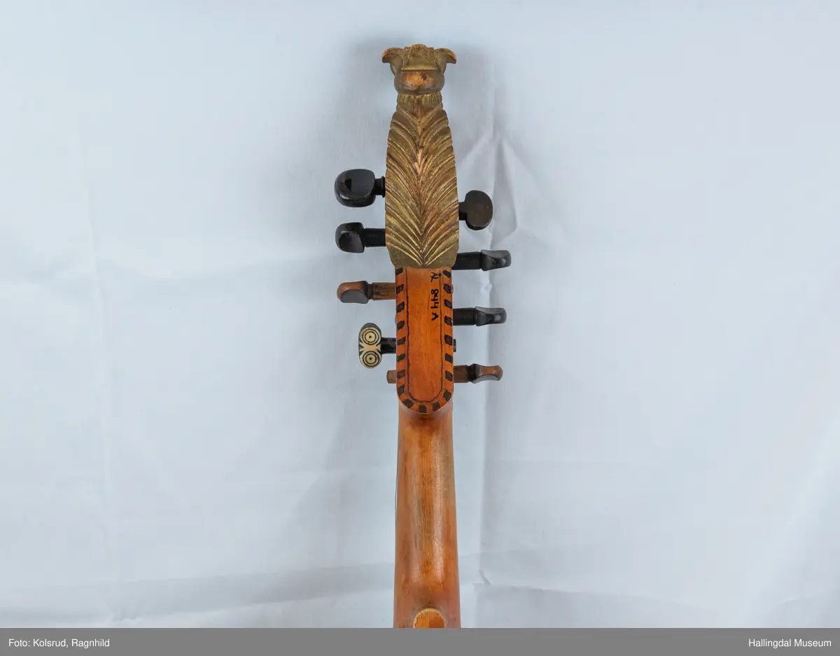 Hardingfele. Fela er litt kuven på midten, og har to S-forma lydhol i loket. Gripebrettet er dekorert med innfelte bitar av perlemor, og det er skore eit dyrehovud med krone i enden av halsen. Fela er dekorert langs kanten, med blomedekor. 