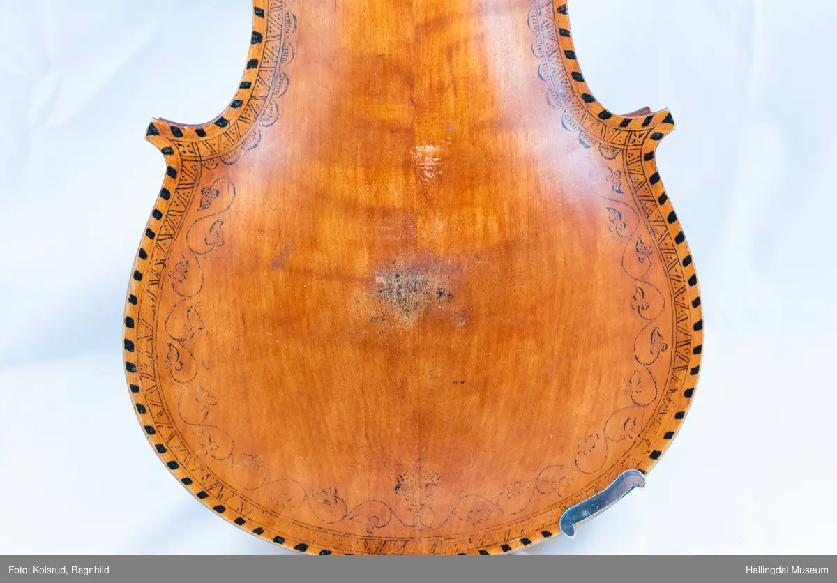 Hardingfele. Fela er litt kuven på midten, og har to S-forma lydhol i loket. Gripebrettet er dekorert med innfelte bitar av perlemor, og det er skore eit dyrehovud med krone i enden av halsen. Fela er dekorert langs kanten, med blomedekor. 