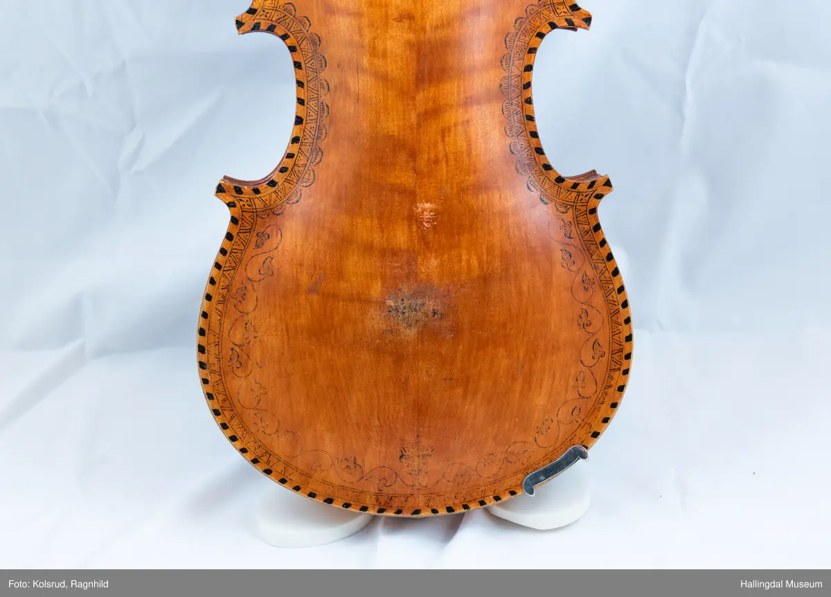 Hardingfele. Fela er litt kuven på midten, og har to S-forma lydhol i loket. Gripebrettet er dekorert med innfelte bitar av perlemor, og det er skore eit dyrehovud med krone i enden av halsen. Fela er dekorert langs kanten, med blomedekor. 