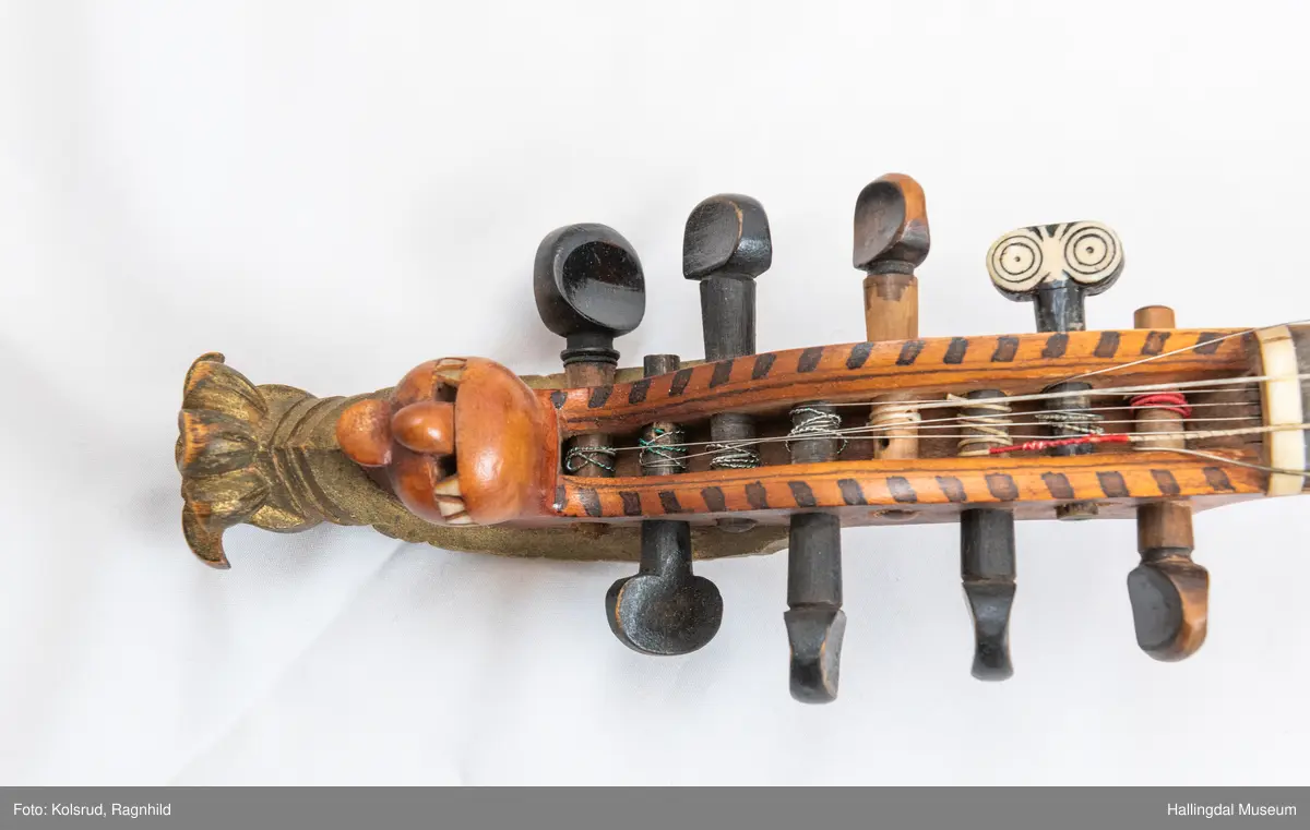 Hardingfele. Fela er litt kuven på midten, og har to S-forma lydhol i loket. Gripebrettet er dekorert med innfelte bitar av perlemor, og det er skore eit dyrehovud med krone i enden av halsen. Fela er dekorert langs kanten, med blomedekor. 