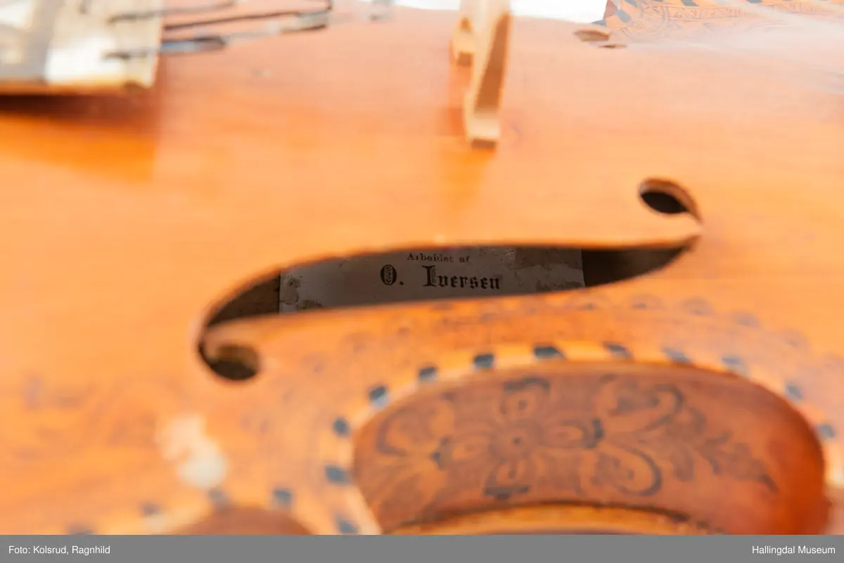 Hardingfele. Fela er litt kuven på midten, og har to S-forma lydhol i loket. Gripebrettet er dekorert med innfelte bitar av perlemor, og det er skore eit dyrehovud med krone i enden av halsen. Fela er dekorert langs kanten, med blomedekor. 