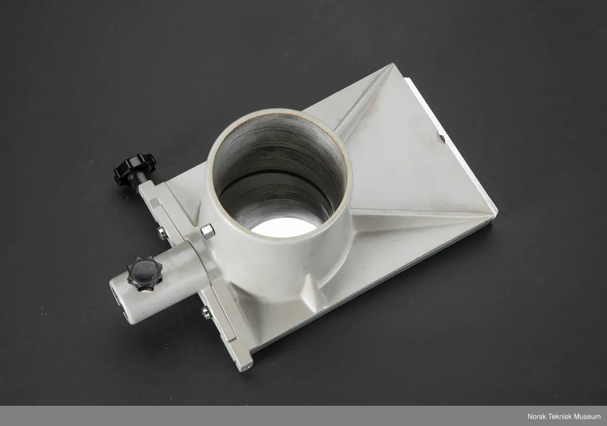 Metallstøpt hoveddel med plate ca A5-størrelse med en solid ring, ca. 10 cm diam. og 10 cm. dyp for innfesting av noe sylindrisk, trolig optikk av noe slag. Hengslet på er en metallramme med speil som kan vippes 90 grader og låses i alle vinkler imellom med en skrue. Påmontert på bunnen er et avtakbart adapter med feste for et stativ, Gjenstanden har ingen merker av noe slag. Tilhører trolig et større instrument eller apparat som er i samlingen.
