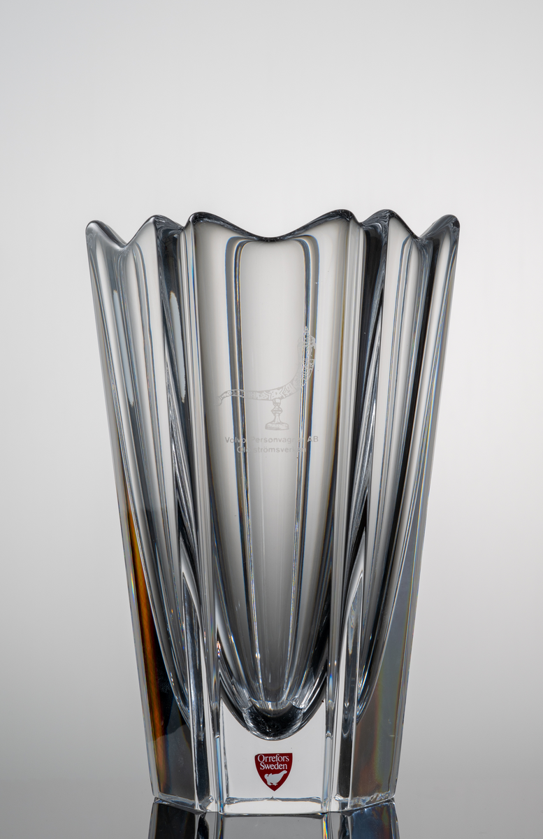 "Corona" Formgivare: Lars Hellsten. Vas i klarglas. Blomkalkformad med sex väggar och profilerad mynning. Avsmalnade mot botten. På kupan ett etsat ymnighetshorn och text ”Volvo Personvagnar AB Olofströmsverken”.