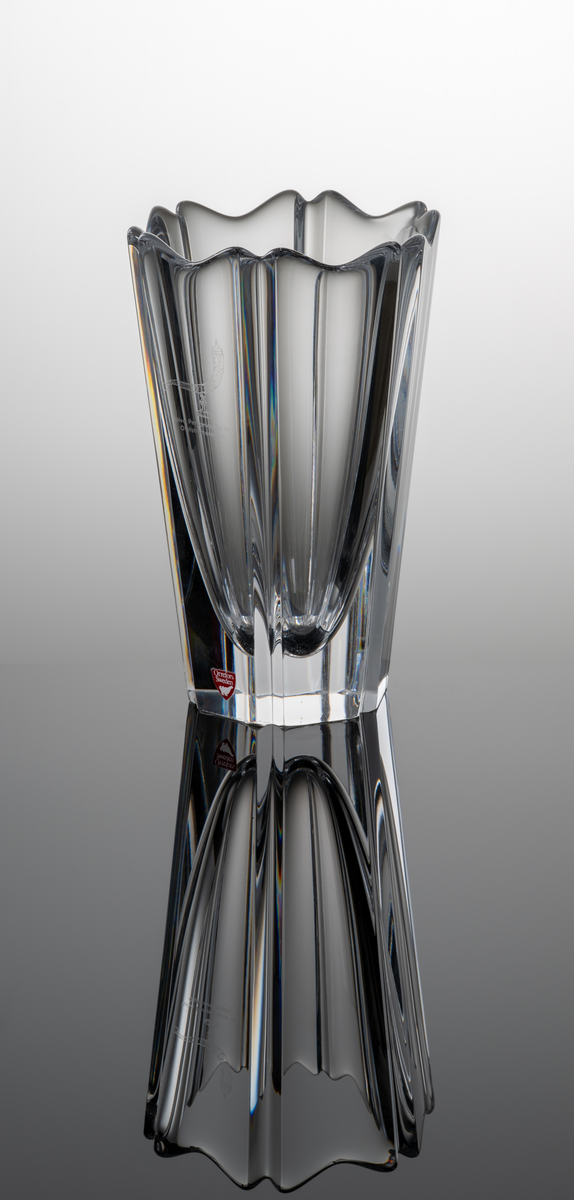 "Corona" Formgivare: Lars Hellsten. Vas i klarglas. Blomkalkformad med sex väggar och profilerad mynning. Avsmalnade mot botten. På kupan ett etsat ymnighetshorn och text ”Volvo Personvagnar AB Olofströmsverken”.