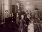 Ronneby Telefonstn år 1932. Telefonsal. Gruppfoto. Vid växelbordet fr.v. ltf E.E. Andersson, lntf I.E. Andersson, ltf Ester Nilsson, ltf Elin Karlsson, Bakre raden fr.v. ltf H. Andersson, rtf K. Rundqvist, rtf J. Olsén, rtf J. Pettersson, ltf A. Hjeltman, tk A. Bergengren, ltf A. Johansson, eltf Ing Andersson.