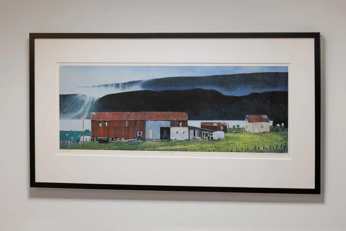 En samling røde og værbitte sjøhus som klynger seg til strandlinjen mot en dypblå fjord og massive, mørke fjell.