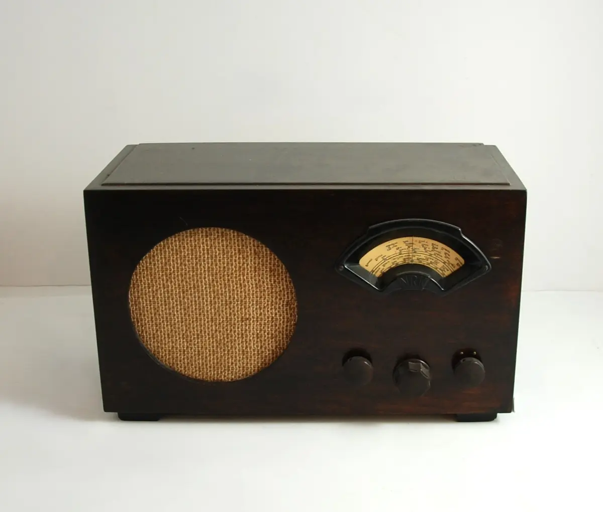 Radio, bordmodell. Rektangulær trekasse av beiset bøk med falset topp og sideplater, samt to lave, rektangulære lister til ben. Fremsiden har et sirkulært hull til høyttaler dekket av en champagnefarget stoff. Stasjonssøkeren har buesegmentformet innramming i sort bakelitt med tre brune bakelittknotter under for tilkobling, av og på, søker og volum. I søkerskivens venstre del står produsentinformasjon. Søkerskiven har lang og mellombølge.