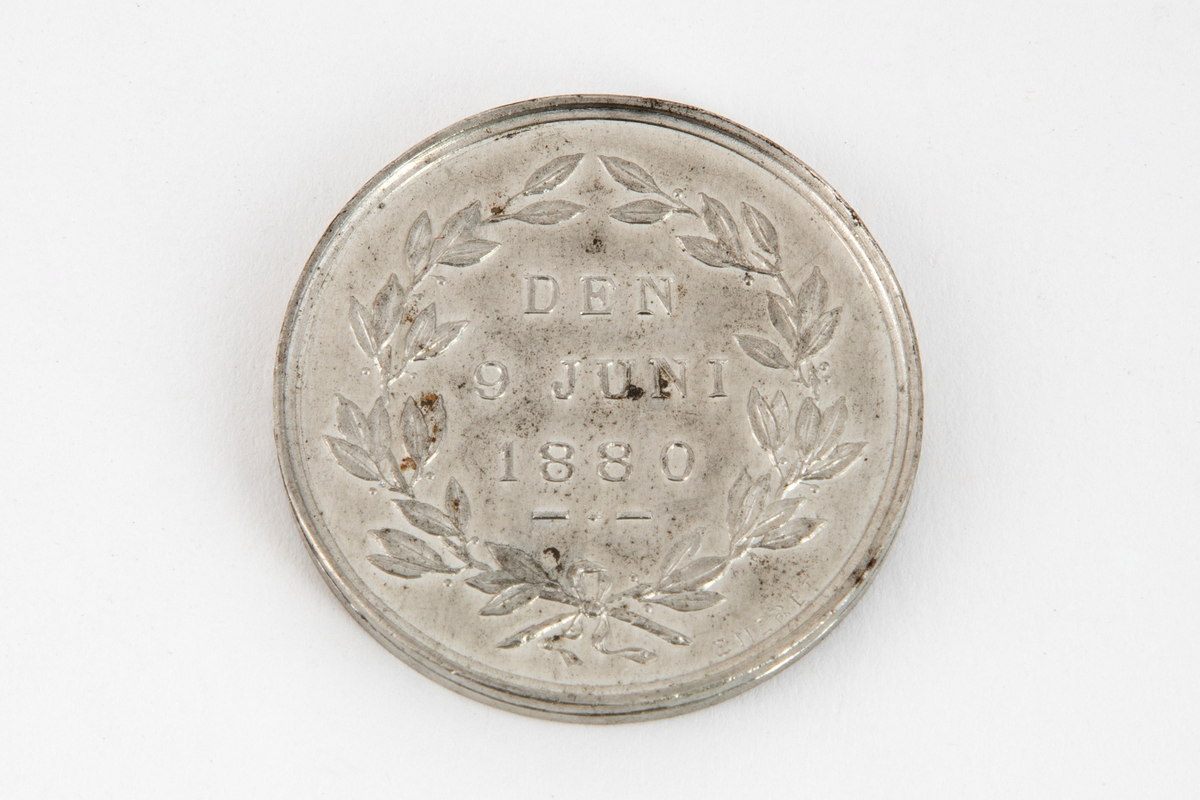 Rund medalje med mannsportett på avers side og laurbærgrener på revers side. Begge sider har omskrift.