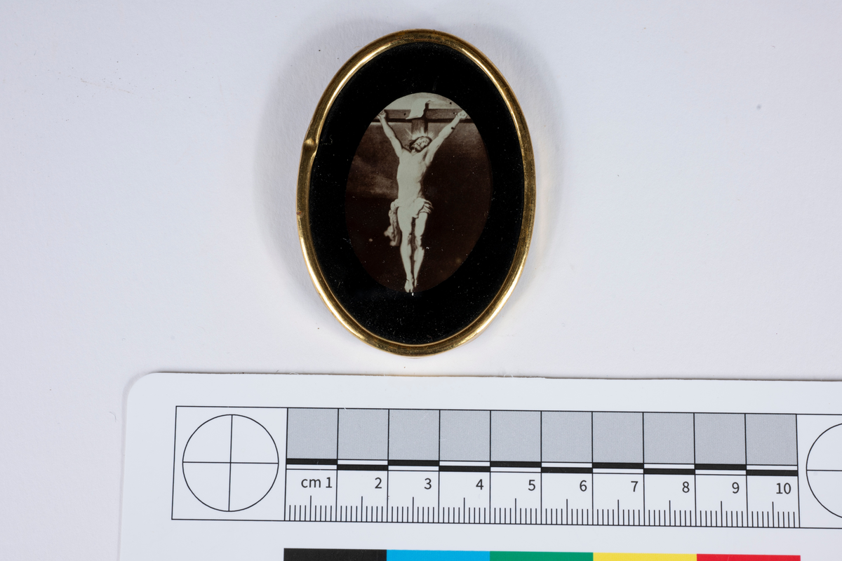 Ovalt fotografi på tavla med Kristi korsfästelse. Svart passepartout. Inglasad med ram av guldfärgad plåt. På baksidan fäste gjort av tygband med en metallring.