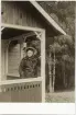 Operasångerskan Christina Nilsson i mörk sidendräkt och ljus hatt. Hon står på verandan till ett enkelt rödmålat hus med 
vita knutar.
Möjl. Loka brunn (?).