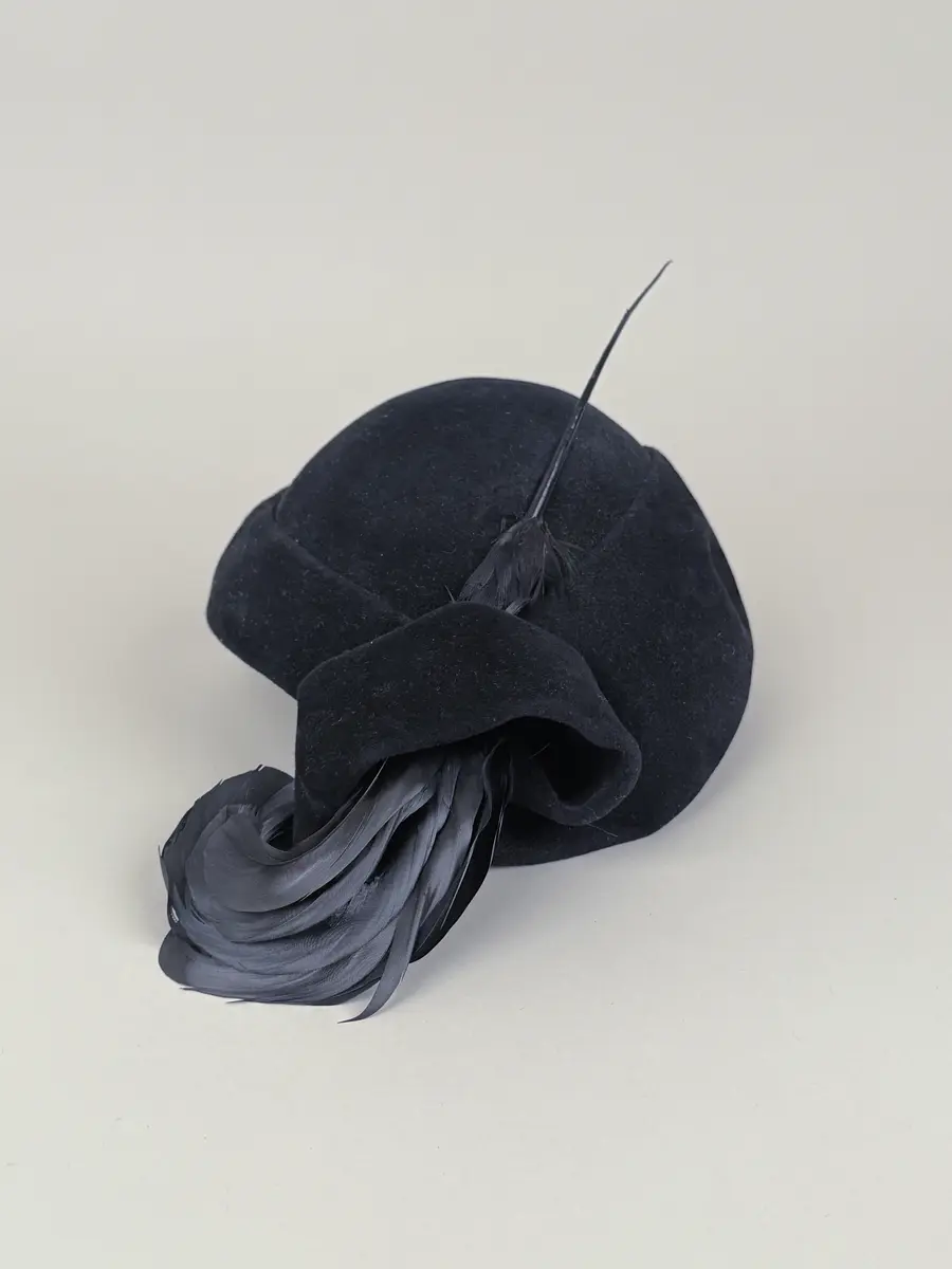 Hatt av moderiktig modell. Asymmetrisk stil med rund kulle och på hattens ena sida är en del av kanten uppvikt och rullad till en spiralform. Genom denna form är en plym monterad. Plymen består av flera rundade och lockade fjärdar sammansatta med en lång spets. Inuti hatten en etikett med invävt firmanamn.