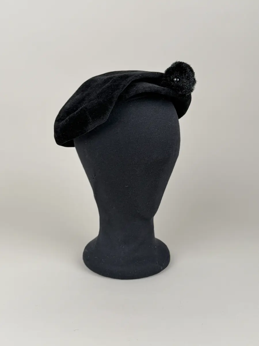 Hatt i baskerliknande form med några veck längs ena sidan. Av svart filtmaterial. Dekorerad med en svart boll monterad med hatt nål med svart runt huvud av plastmaterial.