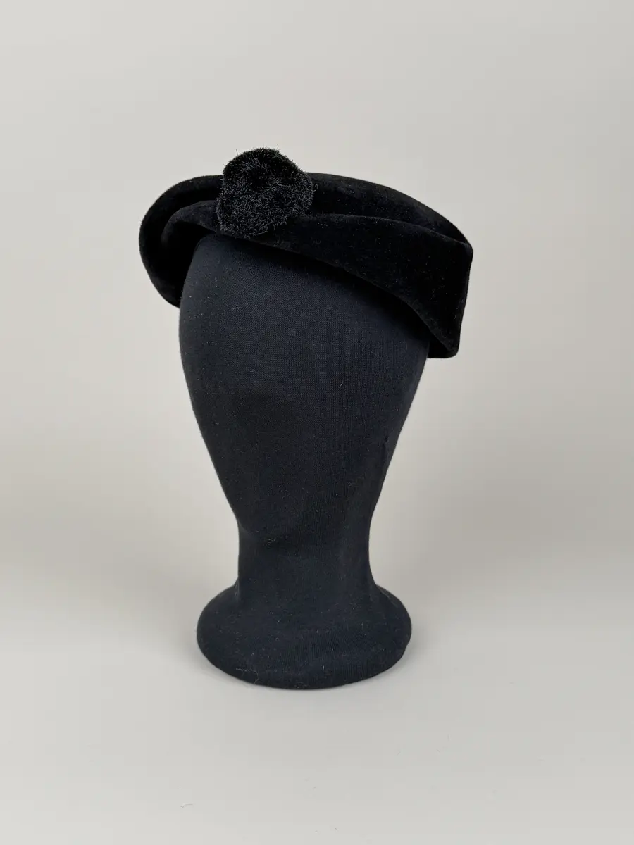 Hatt i baskerliknande form med några veck längs ena sidan. Av svart filtmaterial. Dekorerad med en svart boll monterad med hatt nål med svart runt huvud av plastmaterial.