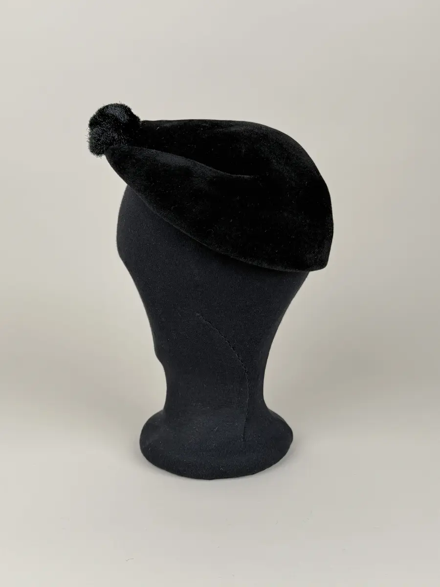 Hatt i baskerliknande form med några veck längs ena sidan. Av svart filtmaterial. Dekorerad med en svart boll monterad med hatt nål med svart runt huvud av plastmaterial.