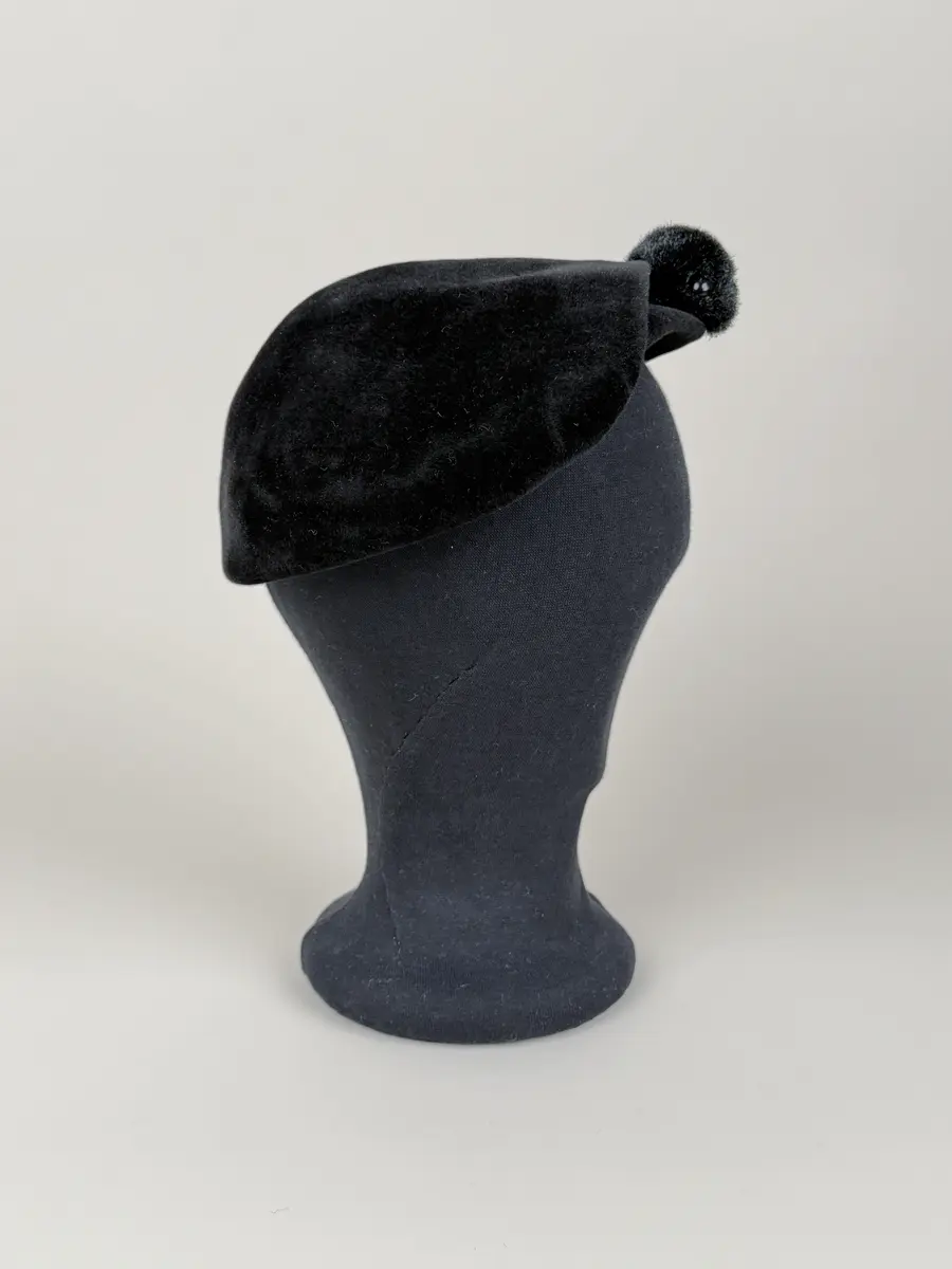 Hatt i baskerliknande form med några veck längs ena sidan. Av svart filtmaterial. Dekorerad med en svart boll monterad med hatt nål med svart runt huvud av plastmaterial.