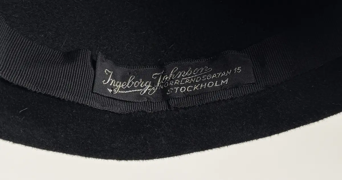 Hatt i baskerliknande form med några veck längs ena sidan. Av svart filtmaterial. Dekorerad med en svart boll monterad med hatt nål med svart runt huvud av plastmaterial.