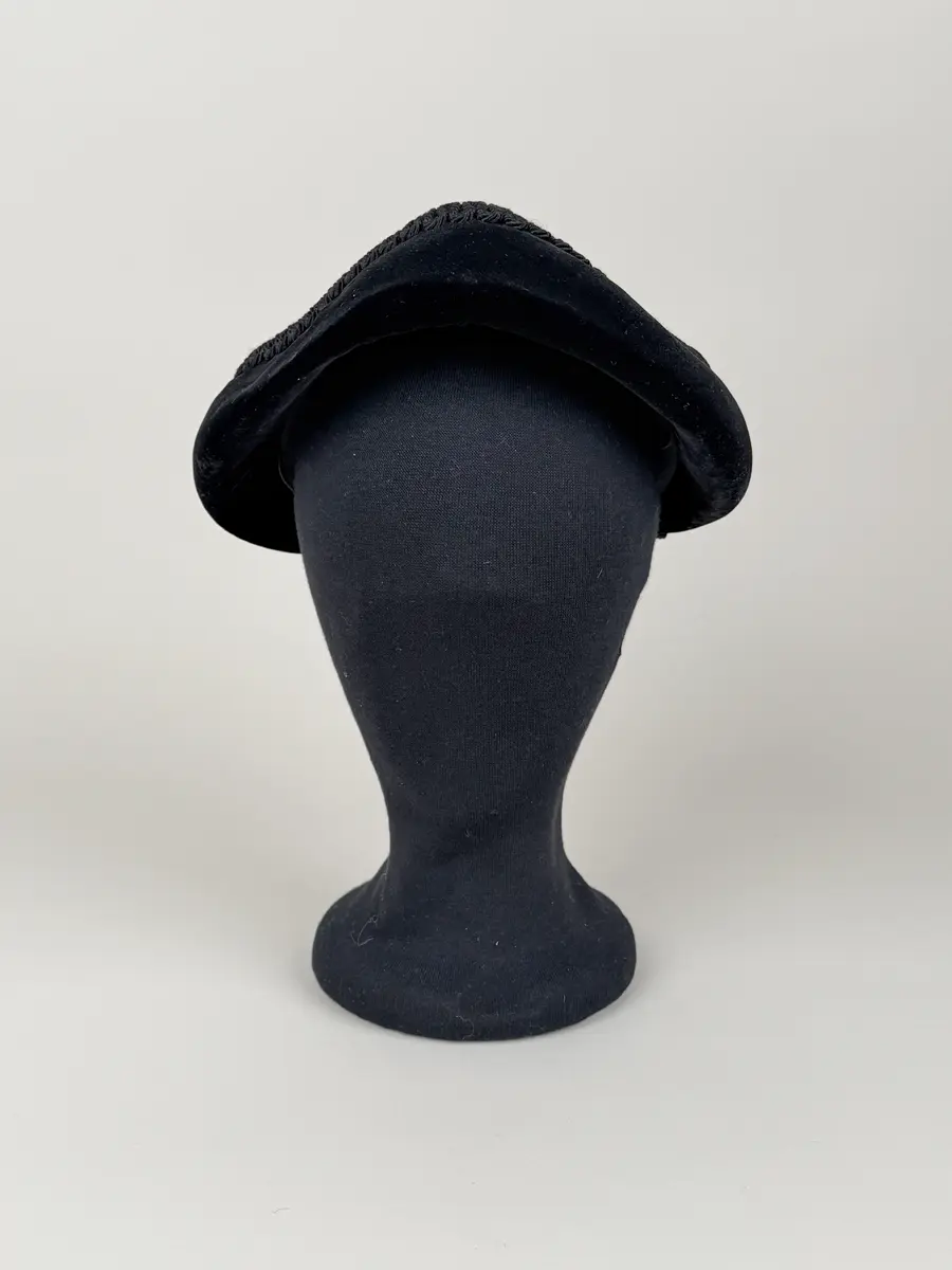 Rund hatt utan brätte. Stomme av styv gles väv av naturfiber klädd med svart sammet. Dekorerad med ett band av en slingrad svart snodd. 